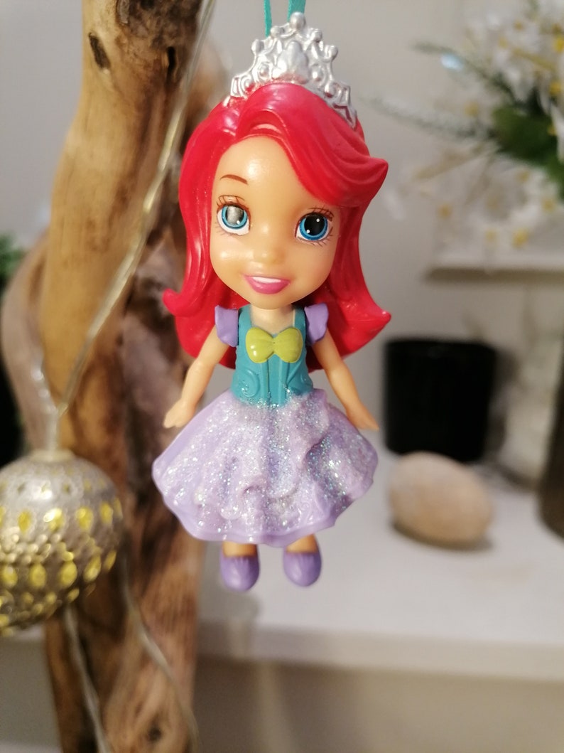 Peut inclure: Une petite poup&eacute;e en plastique d'Ariel de La Petite Sir&egrave;ne portant une robe violette &agrave; paillettes, un corsage turquoise et une couronne argent&eacute;e. Elle a les cheveux roux et les yeux bleus.