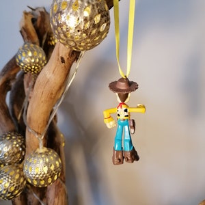 May include: A decorative display featuring a small Woody toy figure hanging from a yellow ribbon. The figure is wearing a brown hat, yellow shirt, blue overalls, and brown boots. The display also includes silver globe lights and a piece of driftwood.