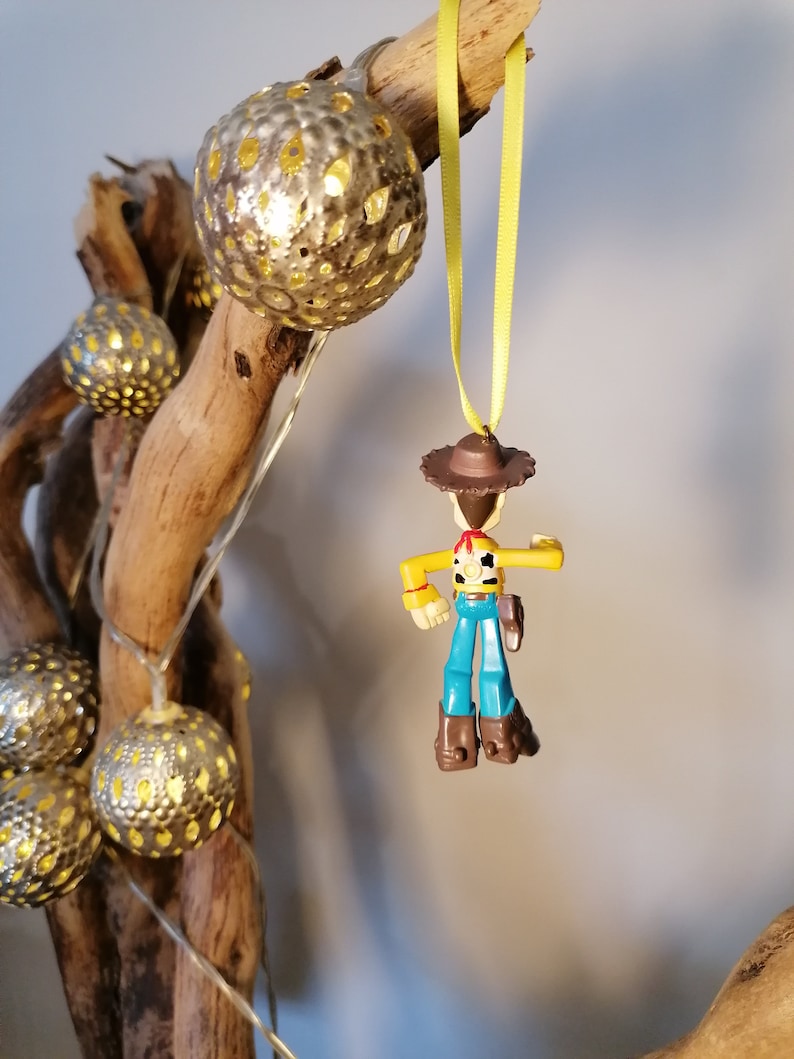 May include: A decorative display featuring a small Woody toy figure hanging from a yellow ribbon. The figure is wearing a brown hat, yellow shirt, blue overalls, and brown boots. The display also includes silver globe lights and a piece of driftwood.