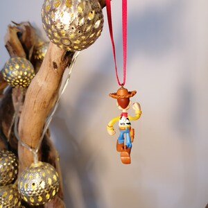 May include: A red string hangs from a branch with a small, brown, plastic figurine of Woody from Toy Story. Woody is wearing his signature cowboy hat, vest, and jeans. He is holding his gun in his right hand and is looking to the right.