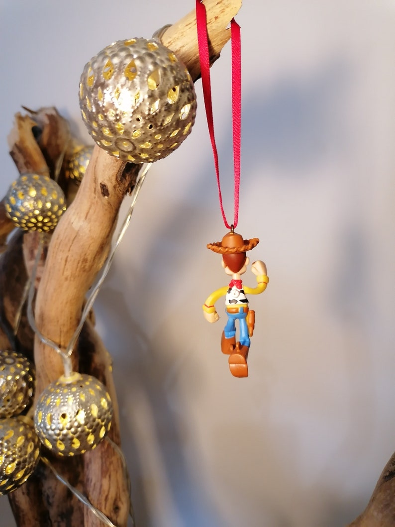 May include: A red string hangs from a branch with a small, brown, plastic figurine of Woody from Toy Story. Woody is wearing his signature cowboy hat, vest, and jeans. He is holding his gun in his right hand and is looking to the right.