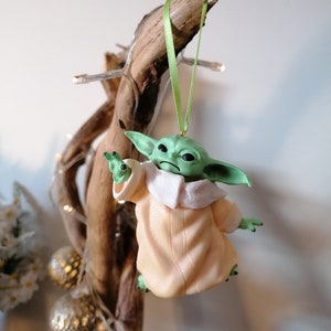 May include: A green and beige Christmas ornament featuring a character from the Star Wars franchise, The Mandalorian. The ornament is hanging from a green ribbon on a branch.