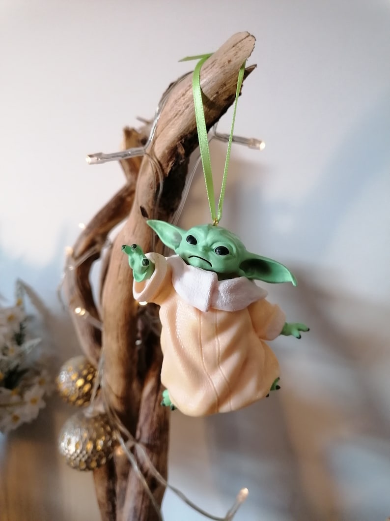 May include: A green and beige Christmas ornament featuring a character from the Star Wars franchise, The Mandalorian. The ornament is hanging from a green ribbon on a branch.
