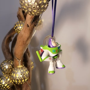 Puede incluir: Un adorno navide&ntilde;o de Buzz Lightyear de pl&aacute;stico morado y blanco colgado de una rama con una cinta morada. El adorno es una peque&ntilde;a figura de pl&aacute;stico de Buzz Lightyear de la franquicia de pel&iacute;culas Toy Story.
