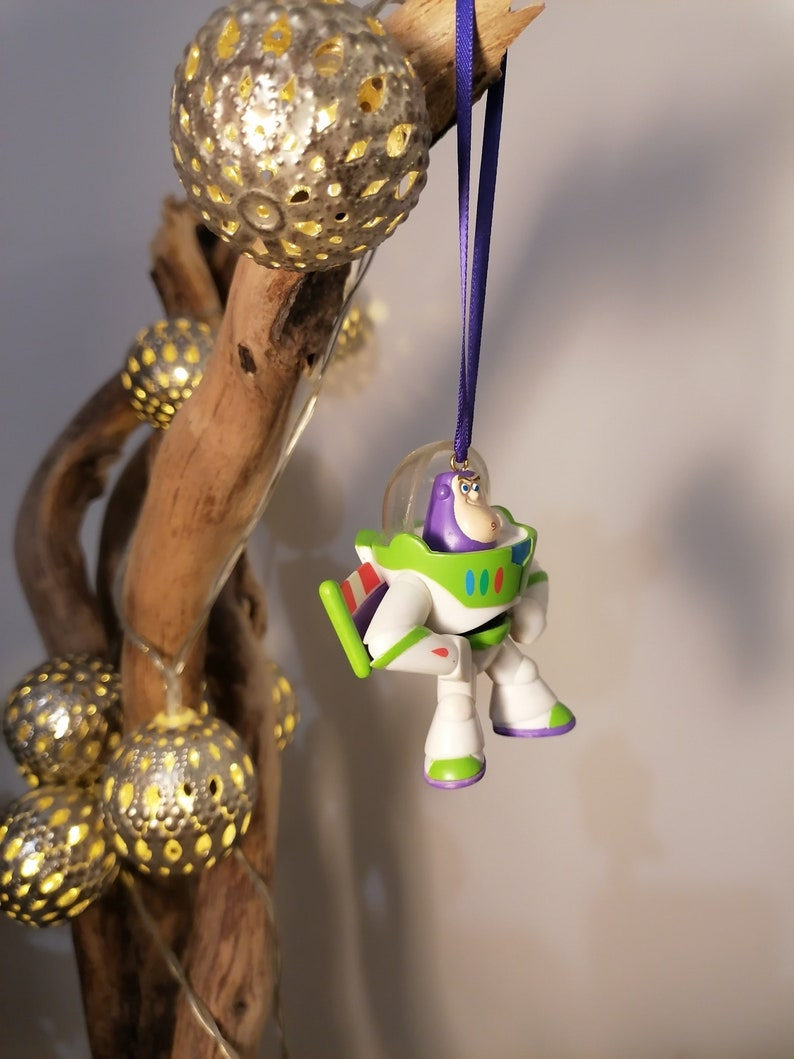 Puede incluir: Un adorno navide&ntilde;o de Buzz Lightyear de pl&aacute;stico morado y blanco colgado de una rama con una cinta morada. El adorno es una peque&ntilde;a figura de pl&aacute;stico de Buzz Lightyear de la franquicia de pel&iacute;culas Toy Story.