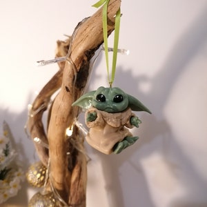 May include: A green and brown Yoda ornament hanging from a branch with a green ribbon. The ornament is a popular character from the Star Wars franchise.