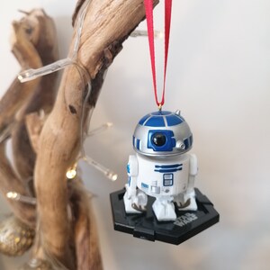 May include: A white and blue Star Wars R2-D2 Christmas ornament hanging from a branch with a red ribbon. The ornament is on a black base with the words "Star Wars" printed on it.