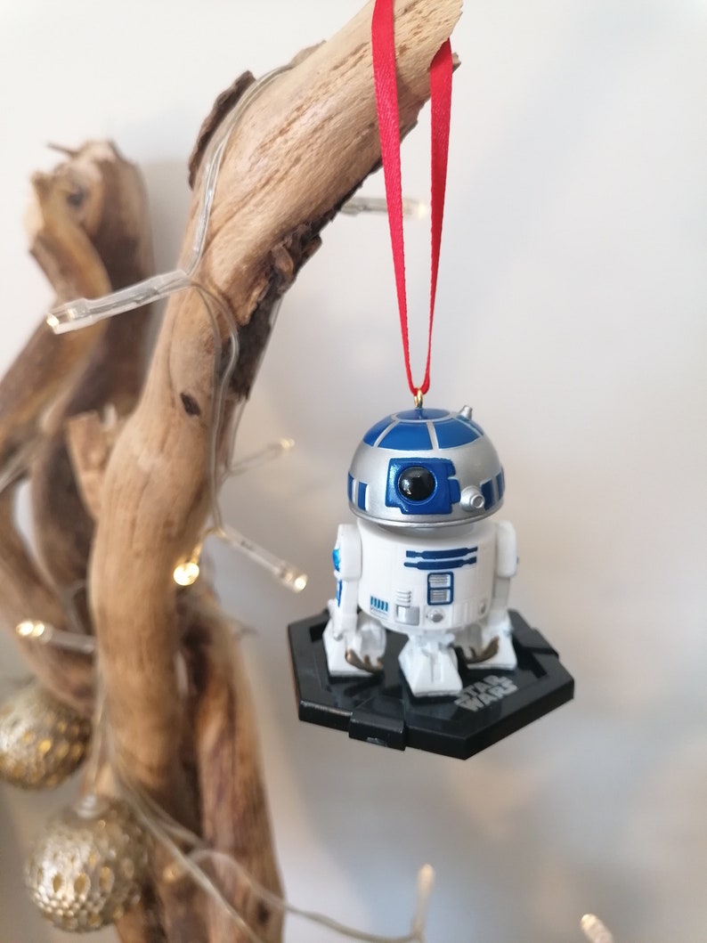 May include: A white and blue Star Wars R2-D2 Christmas ornament hanging from a branch with a red ribbon. The ornament is on a black base with the words "Star Wars" printed on it.