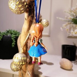 K&ouml;nnte beinhalten: Eine kleine, blaue und rote Plastikfigur eines M&auml;dchens mit braunen Haaren, die an einem blauen Band h&auml;ngt. Die Figur tr&auml;gt ein blaues Kleid mit wei&szlig;en Blumenmustern und roten Akzenten.