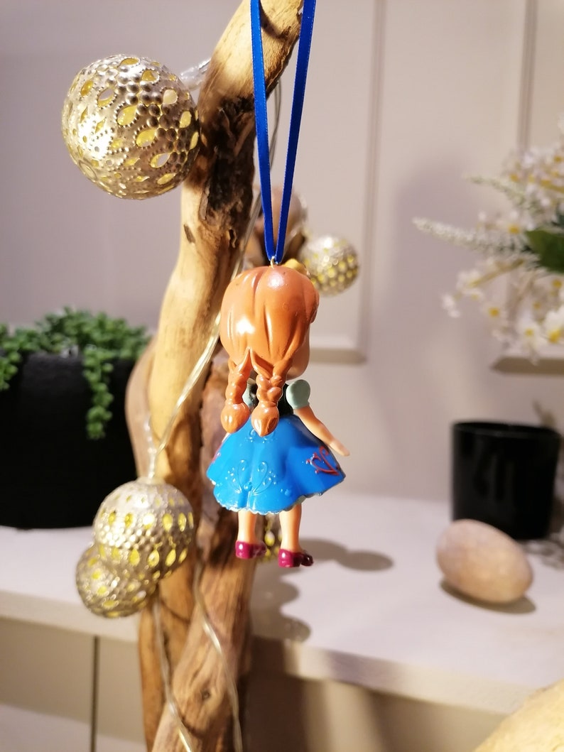 K&ouml;nnte beinhalten: Eine kleine, blaue und rote Plastikfigur eines M&auml;dchens mit braunen Haaren, die an einem blauen Band h&auml;ngt. Die Figur tr&auml;gt ein blaues Kleid mit wei&szlig;en Blumenmustern und roten Akzenten.