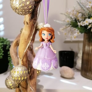 Puede incluir: Un adorno de princesa de color morado y blanco colgando de una rama con una cadena de luces. La muñeca tiene el pelo castaño y una corona.