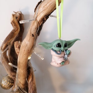 May include: A green and pink Yoda ornament with a green ribbon hanging from a branch. The ornament is holding a silver object in its hand.