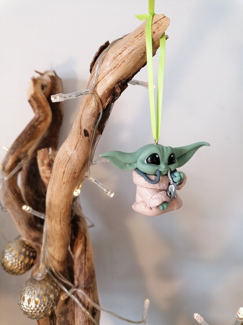 May include: A green and pink Yoda ornament with a green ribbon hanging from a branch. The ornament is holding a silver object in its hand.