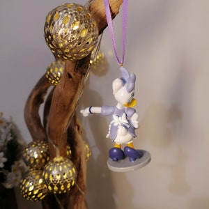 Puede incluir: Una figurita de pl&aacute;stico de un pato de dibujos animados que lleva un vestido azul y un delantal blanco, colgada de una cinta morada en una rama con adornos dorados y plateados.