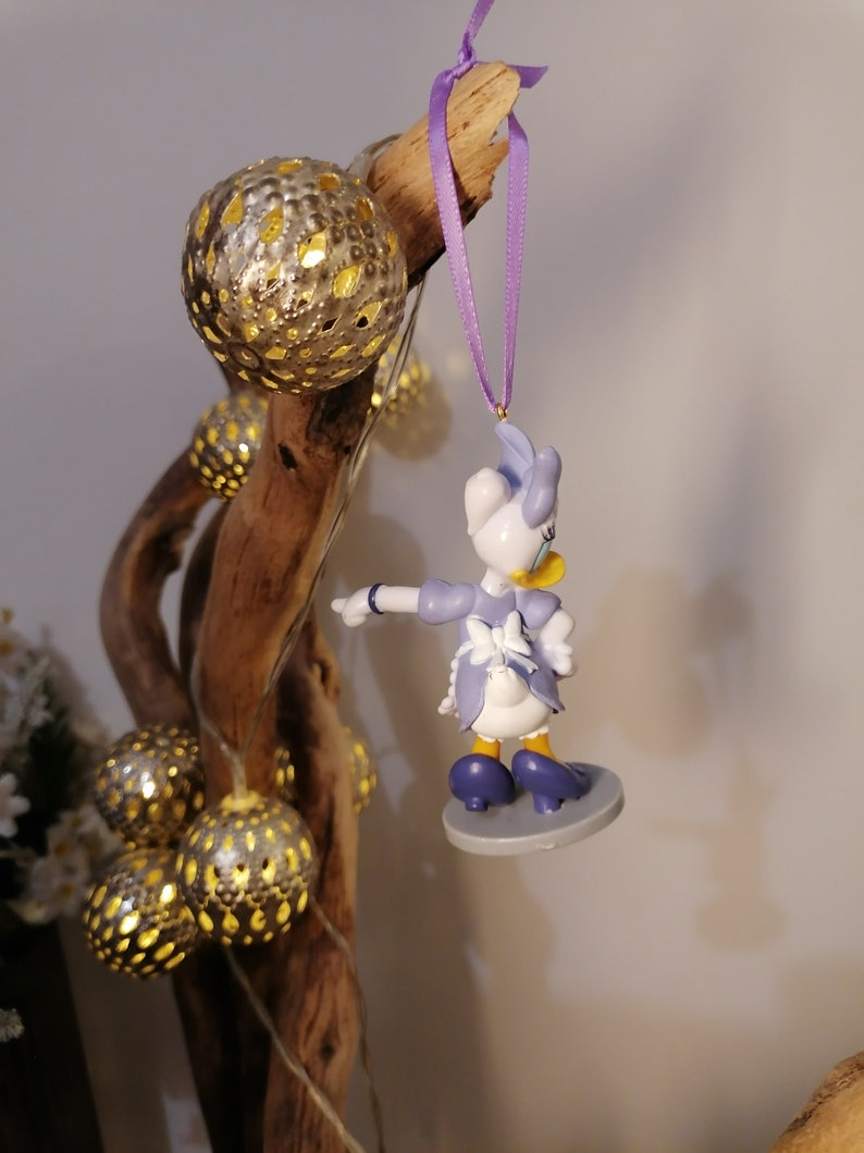 Puede incluir: Una figurita de pl&aacute;stico de un pato de dibujos animados que lleva un vestido azul y un delantal blanco, colgada de una cinta morada en una rama con adornos dorados y plateados.