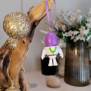 Puede incluir: Un adorno de astronauta de color morado y blanco colgado de una rama con una cinta morada. El adorno est&aacute; hecho de pl&aacute;stico y tiene una visera verde.