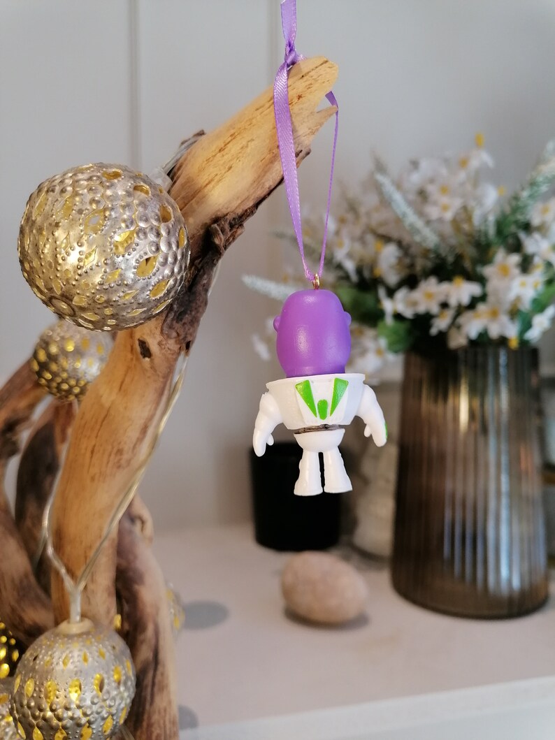Puede incluir: Un adorno de astronauta de color morado y blanco colgado de una rama con una cinta morada. El adorno est&aacute; hecho de pl&aacute;stico y tiene una visera verde.