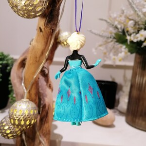 K&ouml;nnte beinhalten: Ein blauer und wei&szlig;er Anh&auml;nger mit einer Plastikpuppe, die ein blaues Kleid mit einem wei&szlig;en Oberteil und einem lila Band um den Hals tr&auml;gt. Die Puppe hat blonde Haare und tr&auml;gt ein blaues und wei&szlig;es Kleid mit einem Schneeflockenmuster.