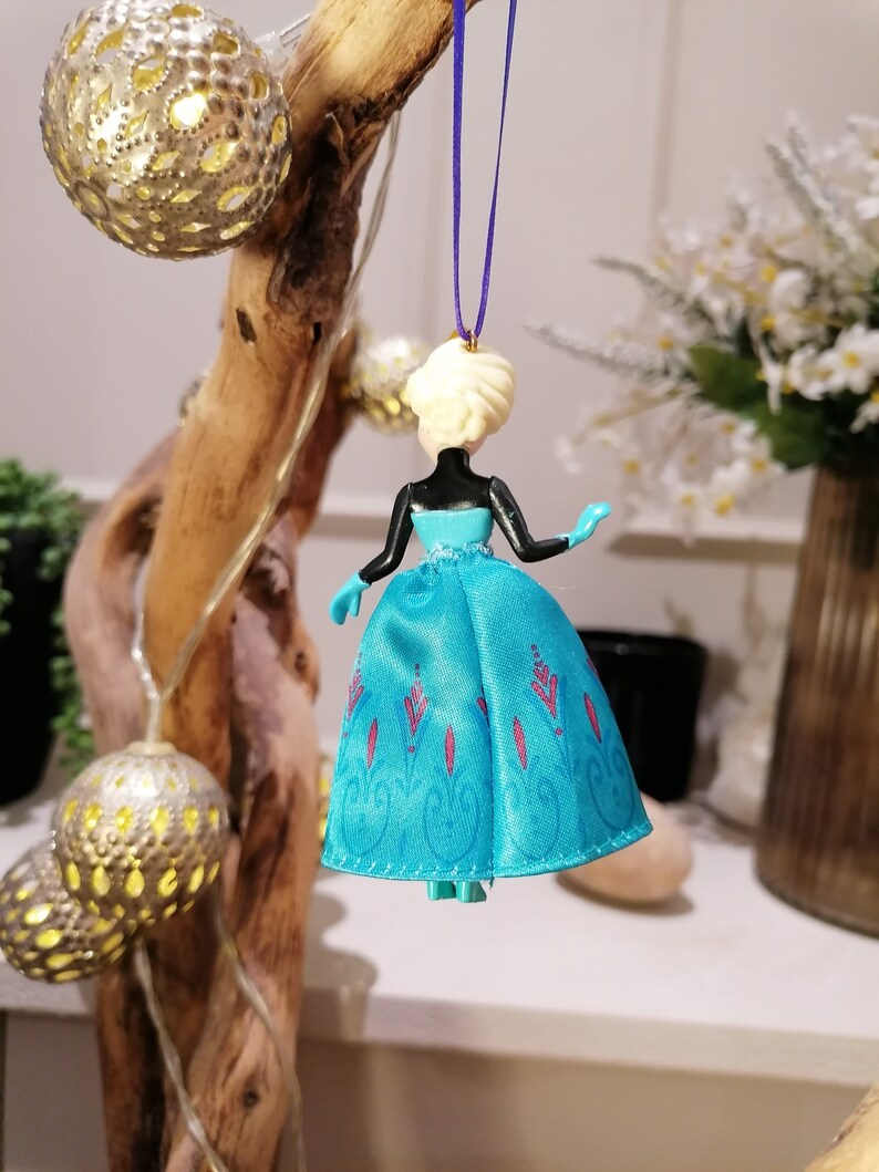 K&ouml;nnte beinhalten: Ein blauer und wei&szlig;er Anh&auml;nger mit einer Plastikpuppe, die ein blaues Kleid mit einem wei&szlig;en Oberteil und einem lila Band um den Hals tr&auml;gt. Die Puppe hat blonde Haare und tr&auml;gt ein blaues und wei&szlig;es Kleid mit einem Schneeflockenmuster.