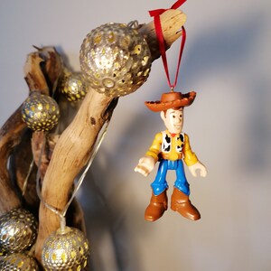 May include: A gold and silver Christmas ornament featuring a plastic figurine of Woody from Toy Story. The ornament is hanging from a branch with a red ribbon.
