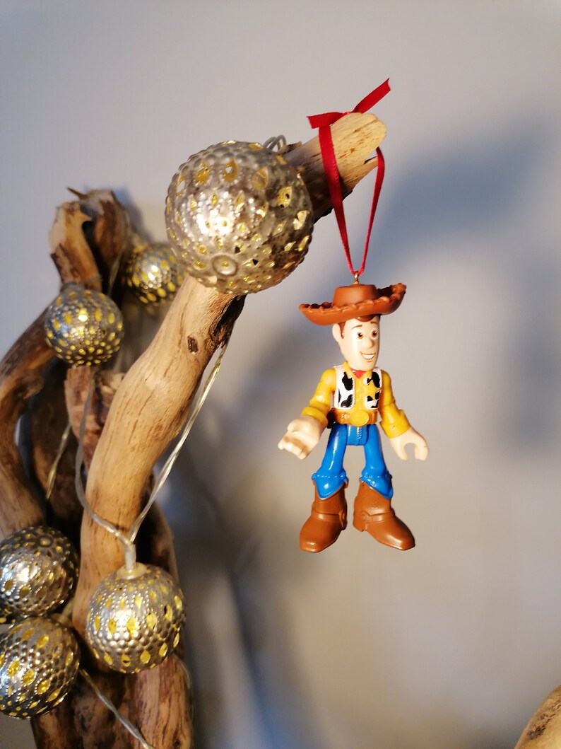May include: A gold and silver Christmas ornament featuring a plastic figurine of Woody from Toy Story. The ornament is hanging from a branch with a red ribbon.