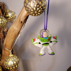 Puede incluir: Un adorno de juguete Buzz Lightyear blanco y verde con detalles morados colgando de una cinta morada en una rama con luces navide&ntilde;as doradas.