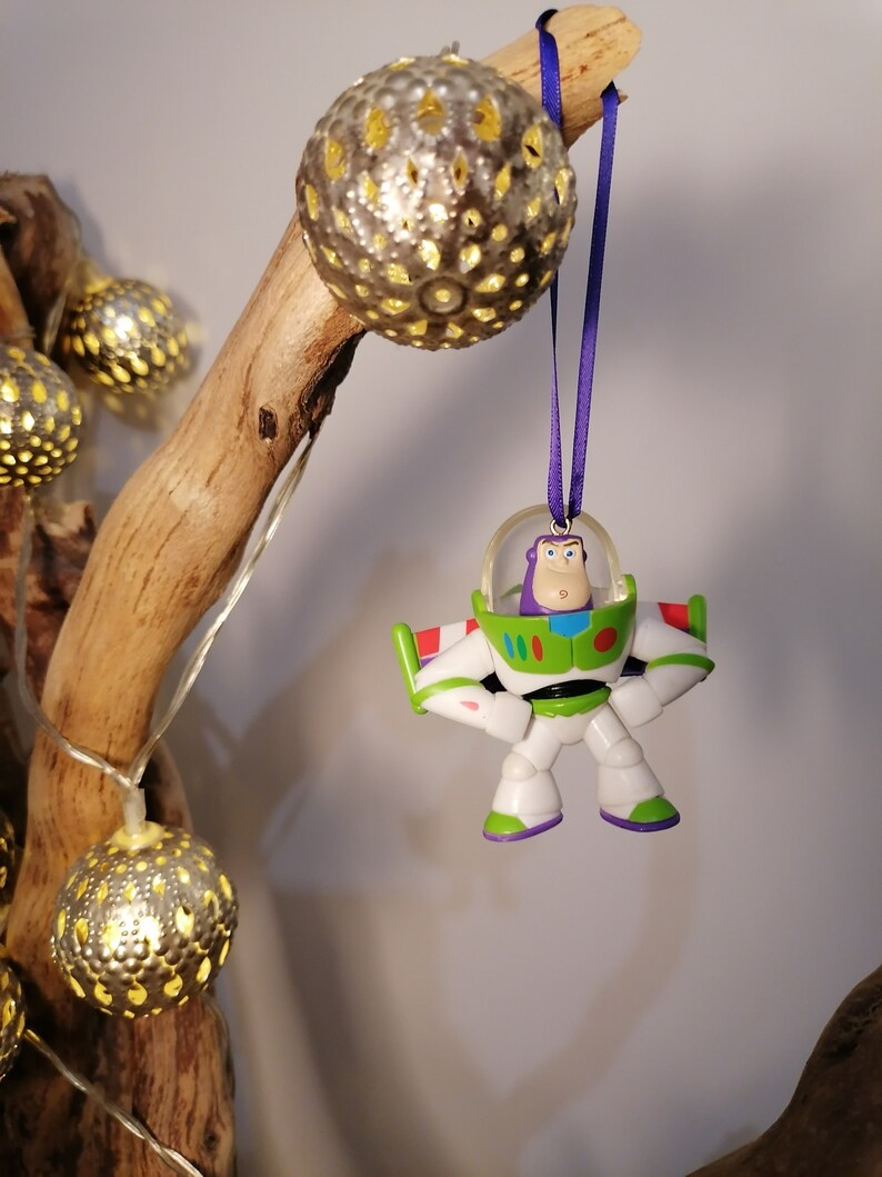 Puede incluir: Un adorno de juguete Buzz Lightyear blanco y verde con detalles morados colgando de una cinta morada en una rama con luces navide&ntilde;as doradas.