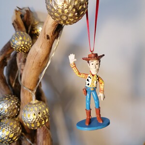 May include: A plastic figurine of Woody from Toy Story, wearing his signature cowboy hat and vest, with a blue base. The figurine is hanging from a red ribbon on a branch with silver, decorative lights.