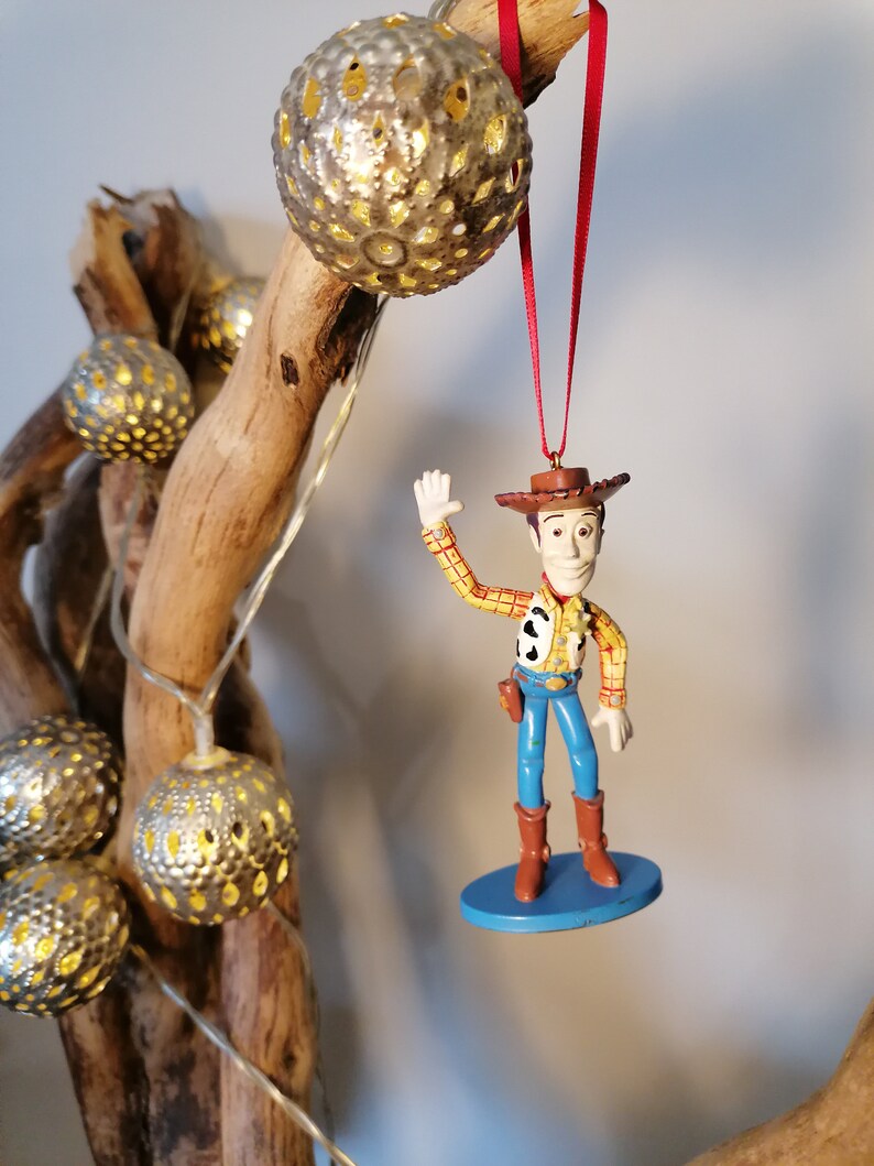 May include: A plastic figurine of Woody from Toy Story, wearing his signature cowboy hat and vest, with a blue base. The figurine is hanging from a red ribbon on a branch with silver, decorative lights.