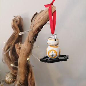May include: A white and orange Star Wars BB-8 character Christmas bauble with a red ribbon hanging from a branch of driftwood.