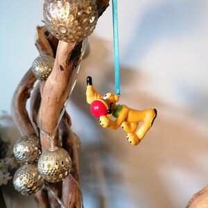 Op de afbeelding: Een geel en bruin cartoonhondje dat aan een blauw lint hangt. De hond houdt een rode bal in zijn bek en hangt aan een tak met zilveren en gouden decoratieve ballen.