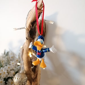 May include: A blue and yellow Donald Duck Christmas ornament with a red ribbon hanging from a piece of driftwood.