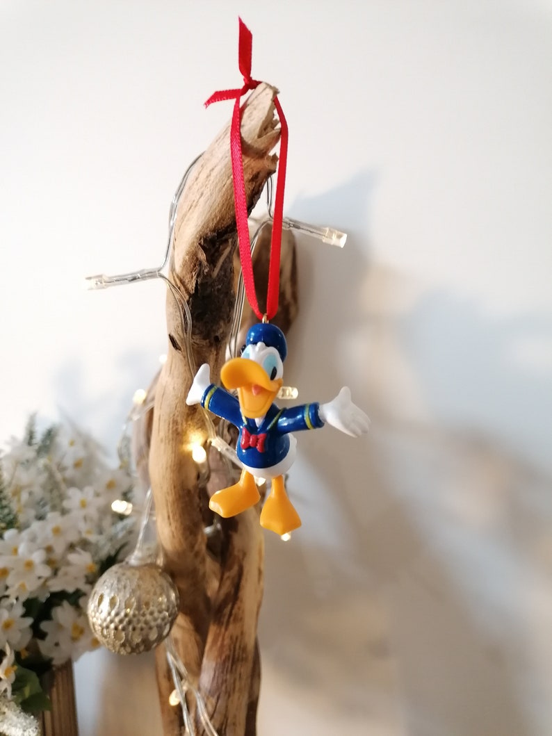 May include: A blue and yellow Donald Duck Christmas ornament with a red ribbon hanging from a piece of driftwood.