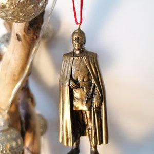 May include: A gold-colored ornament depicting a character from Star Wars wearing a cape and holding a weapon. The ornament is hanging from a red ribbon.