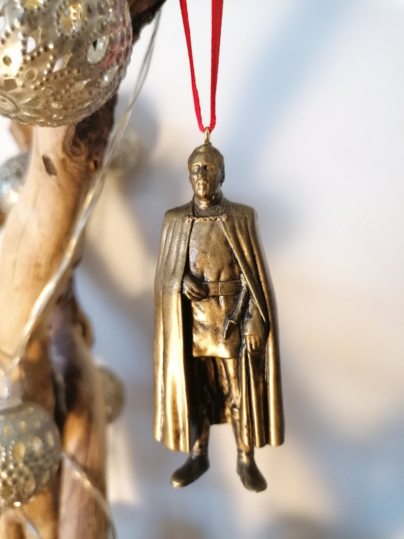 May include: A gold-colored ornament depicting a character from Star Wars wearing a cape and holding a weapon. The ornament is hanging from a red ribbon.