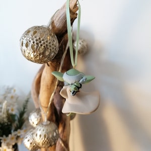 May include: A green and beige Yoda ornament hanging from a branch with a green ribbon. The ornament is a small figurine of Yoda holding a lightsaber.