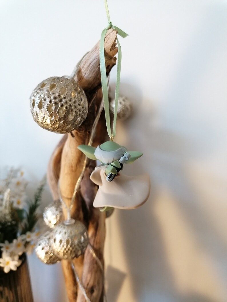 May include: A green and beige Yoda ornament hanging from a branch with a green ribbon. The ornament is a small figurine of Yoda holding a lightsaber.