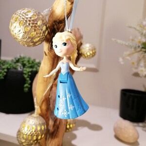 May include: A blue and white plastic ornament of a Disney princess with blonde hair. The ornament is hanging from a branch with string lights.