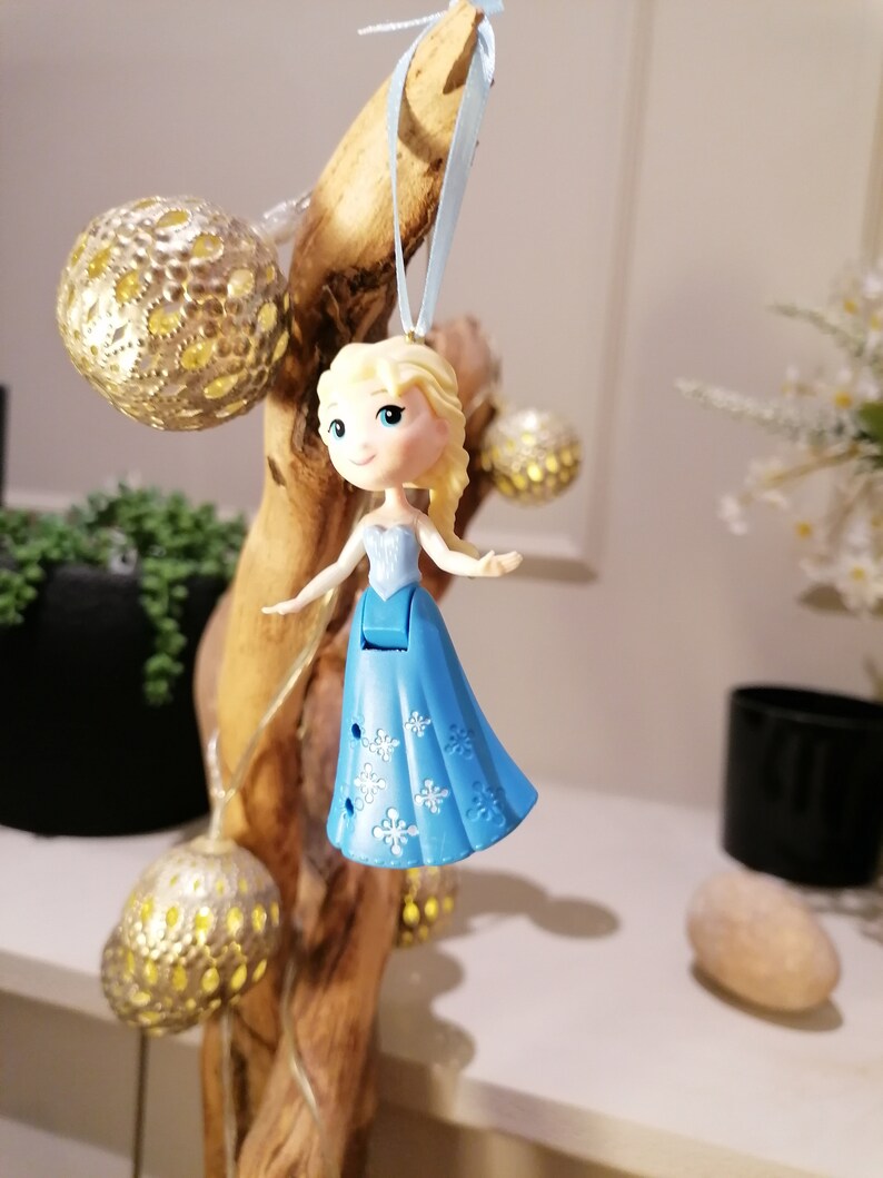 May include: A blue and white plastic ornament of a Disney princess with blonde hair. The ornament is hanging from a branch with string lights.