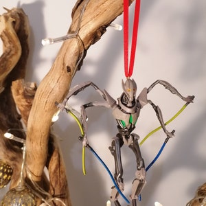 May include: A gray and silver Star Wars General Grievous Christmas ornament with a red ribbon. The ornament features a detailed sculpt of the character with green and blue lightsabers.