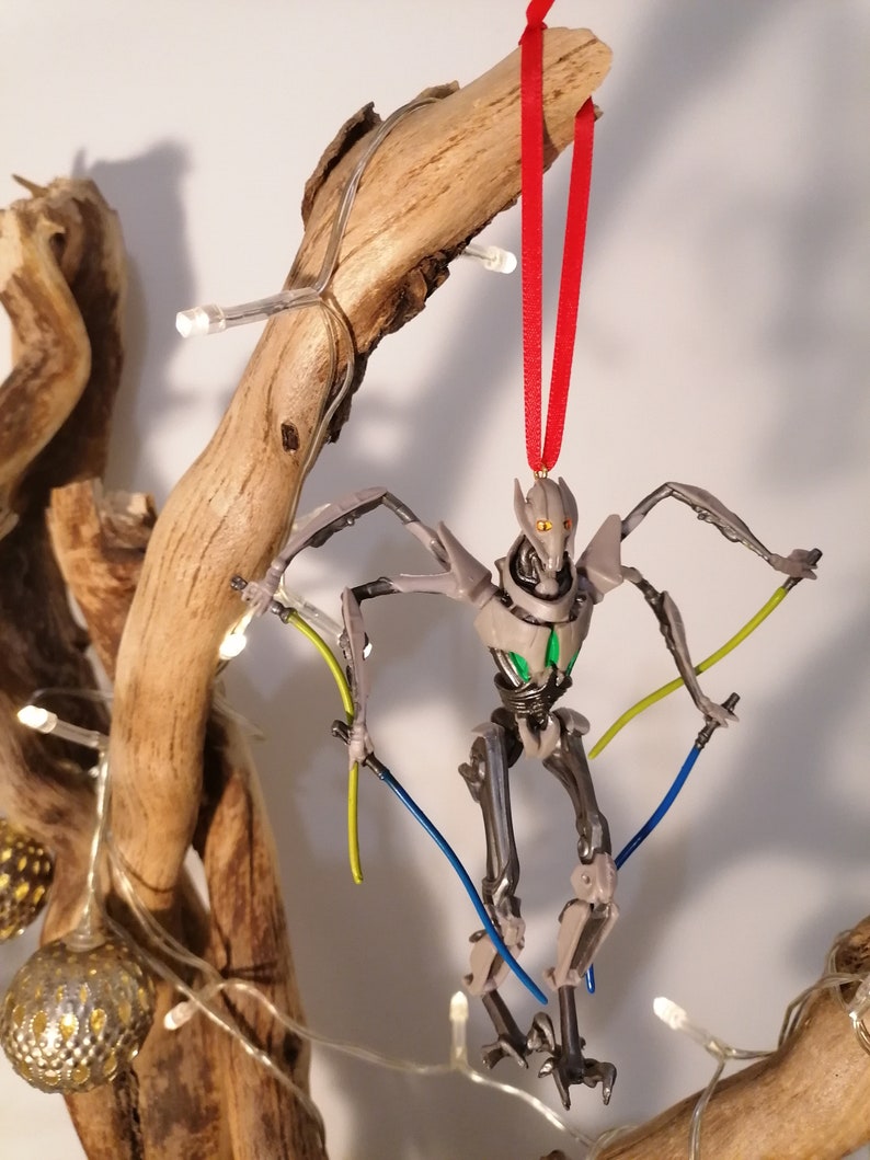 May include: A gray and silver Star Wars General Grievous Christmas ornament with a red ribbon. The ornament features a detailed sculpt of the character with green and blue lightsabers.