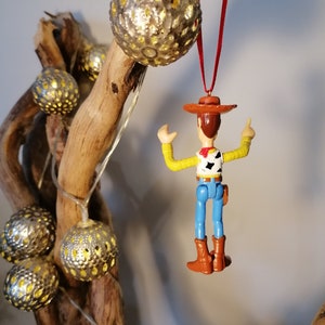 May include: A plastic toy figurine of Woody from Toy Story hanging from a red string. Woody is wearing his signature cowboy hat, vest, and jeans. He is holding his right arm up in the air, and his left arm is bent at the elbow. The figurine is hanging from a branch with gold and white ornaments.