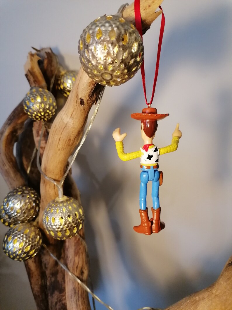 May include: A plastic toy figurine of Woody from Toy Story hanging from a red string. Woody is wearing his signature cowboy hat, vest, and jeans. He is holding his right arm up in the air, and his left arm is bent at the elbow. The figurine is hanging from a branch with gold and white ornaments.