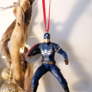 May include: A blue and red Captain America Christmas ornament with a red ribbon. The ornament features a detailed figure of Captain America holding his shield.
