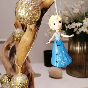 May include: A blue and white ornament featuring a cartoon character with blonde hair and a blue dress. The ornament is hanging from a branch with string lights and gold ornaments.