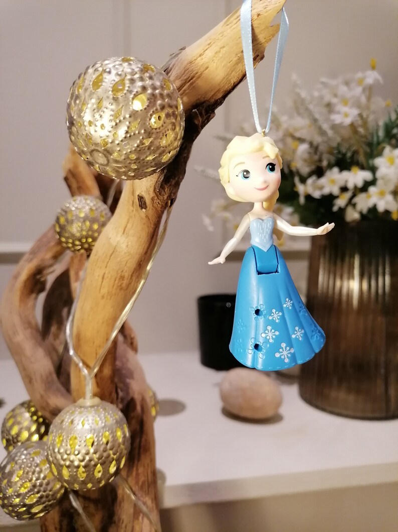 May include: A blue and white ornament featuring a cartoon character with blonde hair and a blue dress. The ornament is hanging from a branch with string lights and gold ornaments.