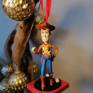 Puede incluir: Un adorno de juguete de vaquero rojo y blanco colgado de una cinta roja. El juguete lleva un sombrero marr&oacute;n, una camisa roja y amarilla, pantalones vaqueros azules y botas marrones. El juguete est&aacute; de pie sobre una plataforma roja y negra.