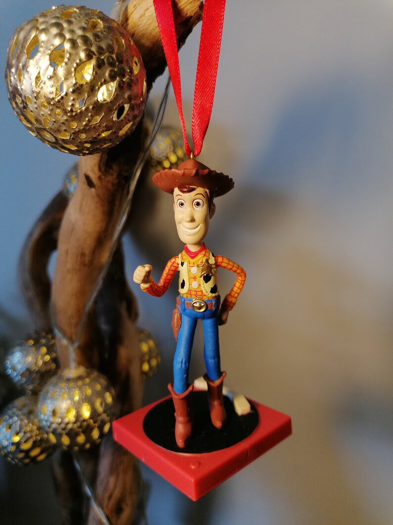 Puede incluir: Un adorno de juguete de vaquero rojo y blanco colgado de una cinta roja. El juguete lleva un sombrero marr&oacute;n, una camisa roja y amarilla, pantalones vaqueros azules y botas marrones. El juguete est&aacute; de pie sobre una plataforma roja y negra.