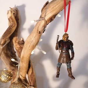 May include: A brown and gold action figure of a warrior wearing armor and holding a weapon, hanging from a branch with a red ribbon. The figure is a Christmas ornament.