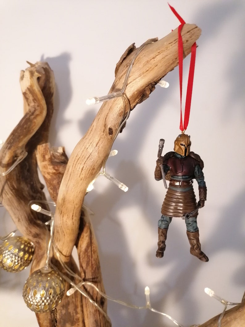 May include: A brown and gold action figure of a warrior wearing armor and holding a weapon, hanging from a branch with a red ribbon. The figure is a Christmas ornament.